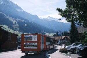 A red bus travels through a picturesque Swiss village surrounded by majestic mountains.